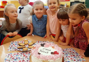 Dziewczynki stoją przy stole. Na stole stoi duży tort z napisem "Dzień kobiet".
